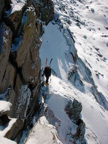 Homme encordé à une paroi montagneuse, portant des skis sur son dos, se déplaçant avec des crampons dans la neige et franchissant des blocs rocheux, dans une activité mêlant alpinisme et ski. Homme encordé à une paroi montagneuse, portant des skis sur son dos, se déplaçant avec des crampons dans la neige et franchissant des blocs rocheux, dans une activité mêlant alpinisme et ski.